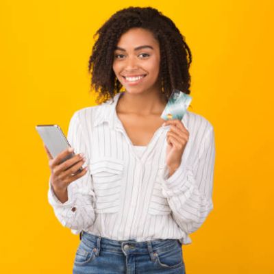 Online Store. Happy black girl holding credit card and mobile phone, ready to buy something. Copyspace, yellow studio